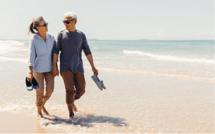 An older couple holding hands and their shoes as they walk barefoot in the water along a sandy beach.