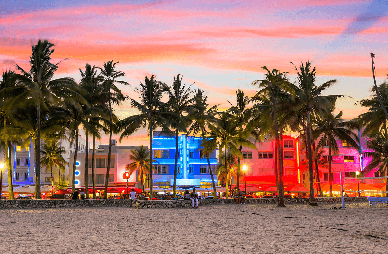 A sandy beach and palm trees in front of colorfully lit buildings along Miami Beach in Florida.