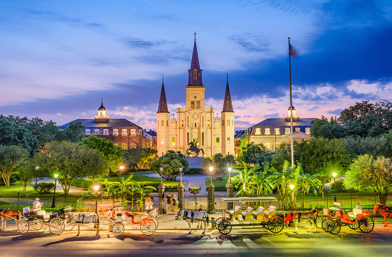 Jackson Square and the St. Louis Cathedral in New Orleans’ French Quarter at dusk with horse-drawn carriages out front.
