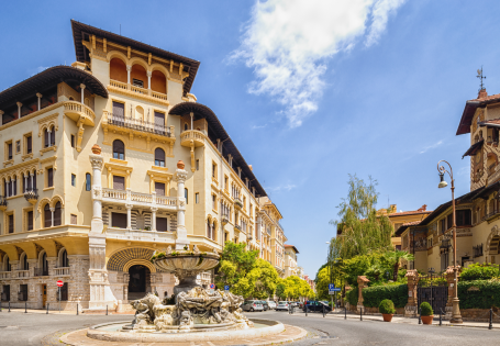 A frontal exterior view of a beautifully cream colored hotel with a fountain in front of it