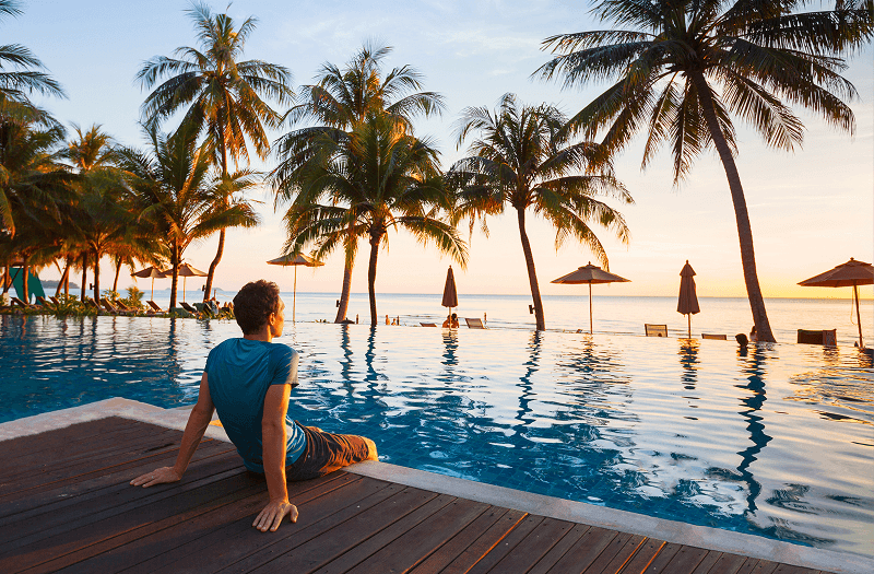 View of a person sitting on the edge of a pool, with palm trees and a sunset sky in the background