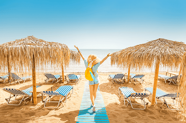 A woman walking in the sand towards the beach with her arms out reached in the sunlight with beach chairs around and a cabana around her.