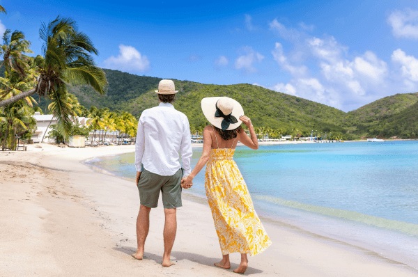 Man and woman walking along white sandy beach with lush green hills in view