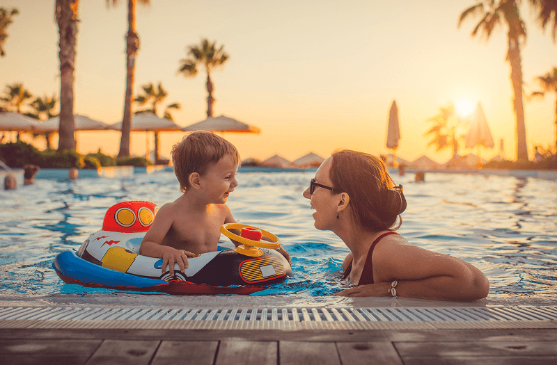 A mother in a swimming pool smiling at her young child, who is sitting in a colorful, toy-like inflatable boat float during a warm sunset.