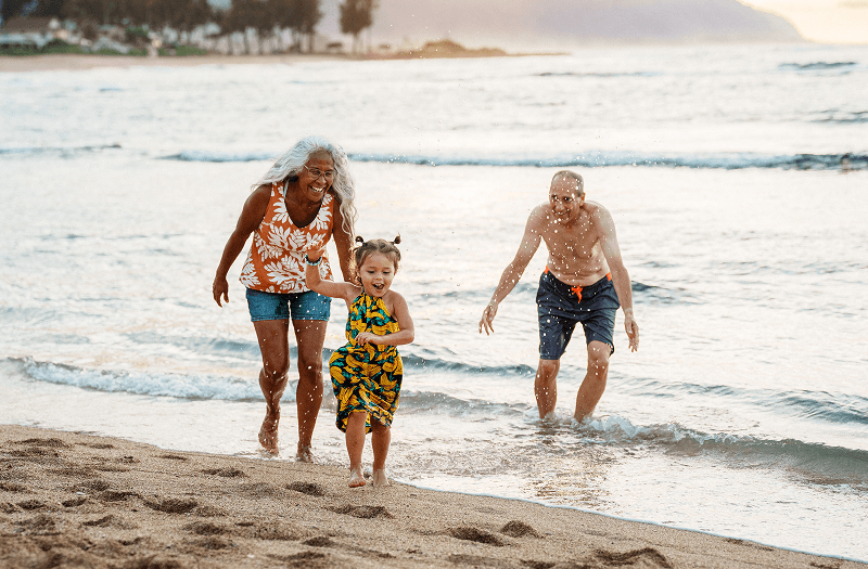 A happy family, including a grandmother, grandfather, and a young girl in a yellow dress, splashing and running together along a sandy ocean beach.