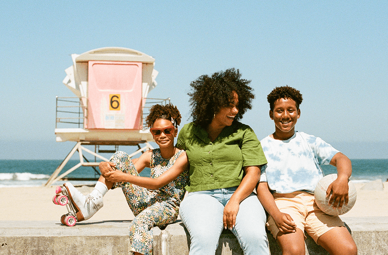 A smiling mom, son holding a basketball, and daughter with roller skates sitting on a ledge in front of a beach on a clear day.