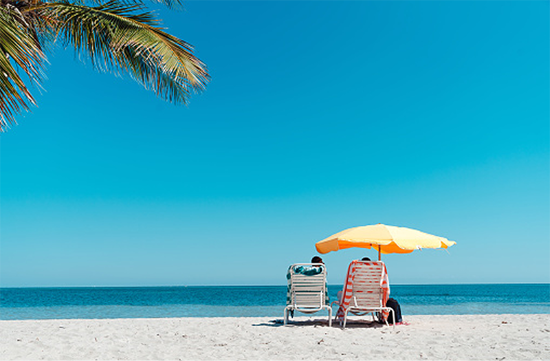 Two people sitting in lounge chairs under a bright yellow umbrella on a white sandy beach, gazing at the ocean.