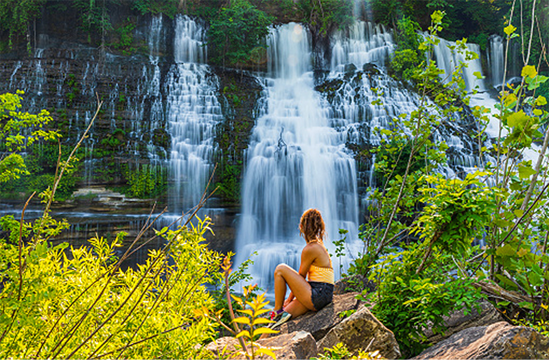 A woman sitting on a boulder, admiring a large, multi-tiered waterfall surrounded by dense green forest.