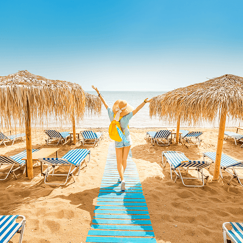 A young lady walking towards the ocean on the beach with her arms outstetched. Surrounding her are beach chairs and cabanas
