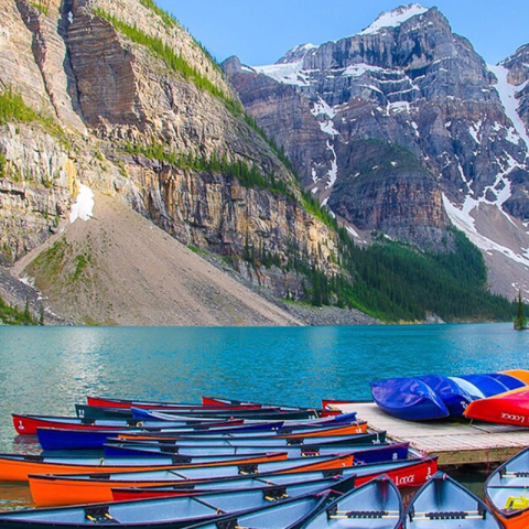 A row of colorful canoes rests at the dock of a tranquil mountain lake, with majestic mountains in the background.  