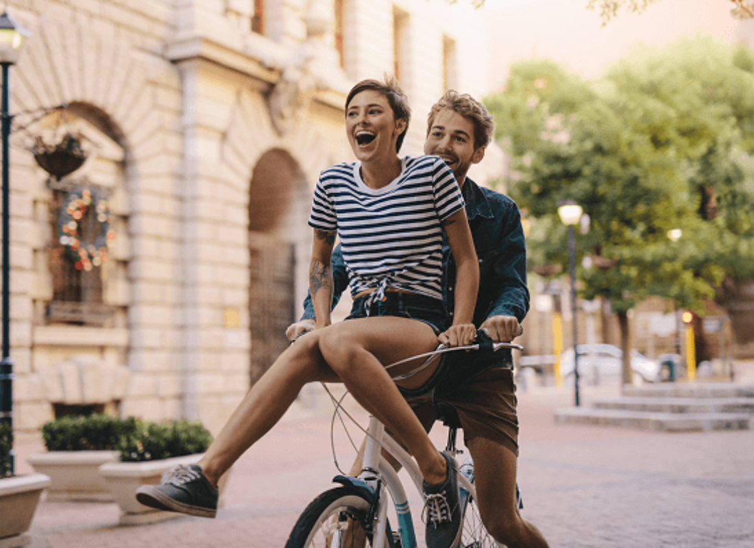 A smiling man riding a bike down a beautiful city road while an excited woman sits on the handlebars.