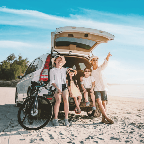 A family of four sits in the open trunk of a SUV parked on a sunny beach, with the father pointing toward the horizon.