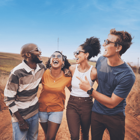 A group of four friends wearing sunglasses laugh and walk together along a dirt road under a blue sky.