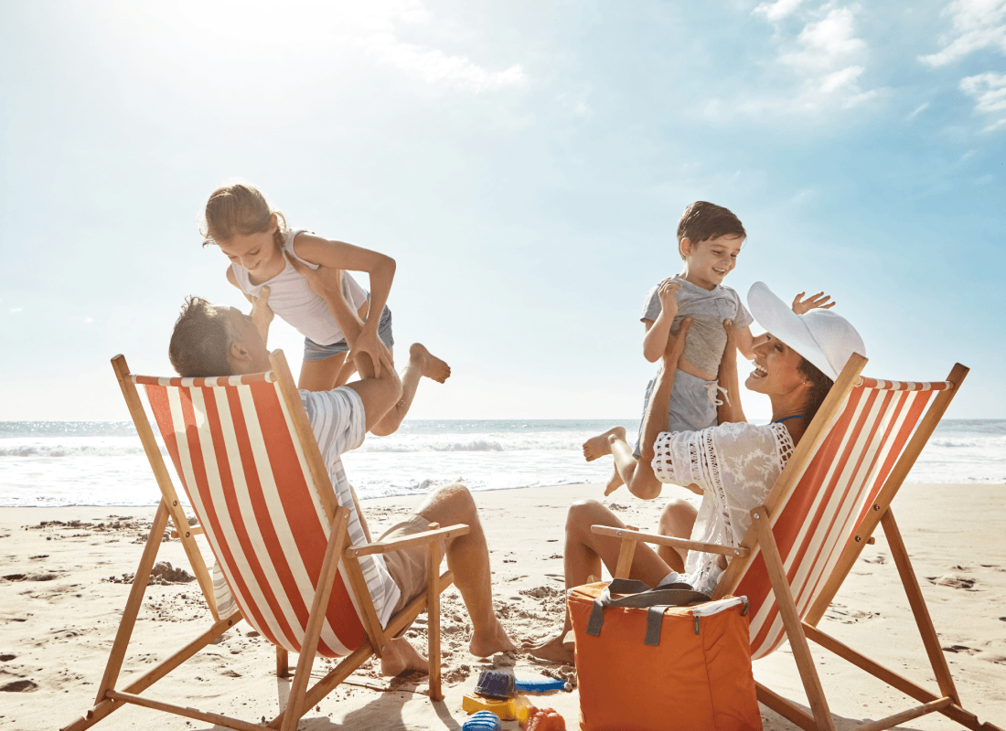 Parents sitting in striped lounge chairs on a sunny beach, lifting their two young children into the air.