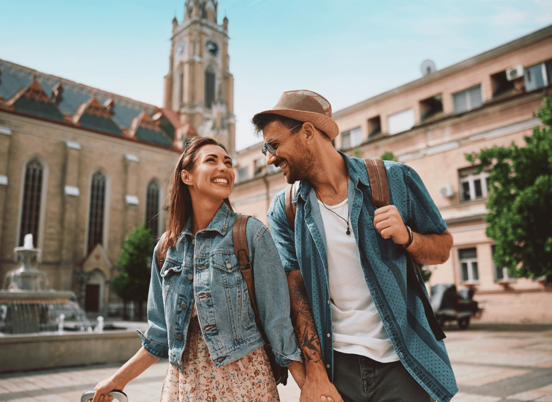 A smiling couple holding hands and pulling luggage walks through a European city square in front of a historic cathedral.