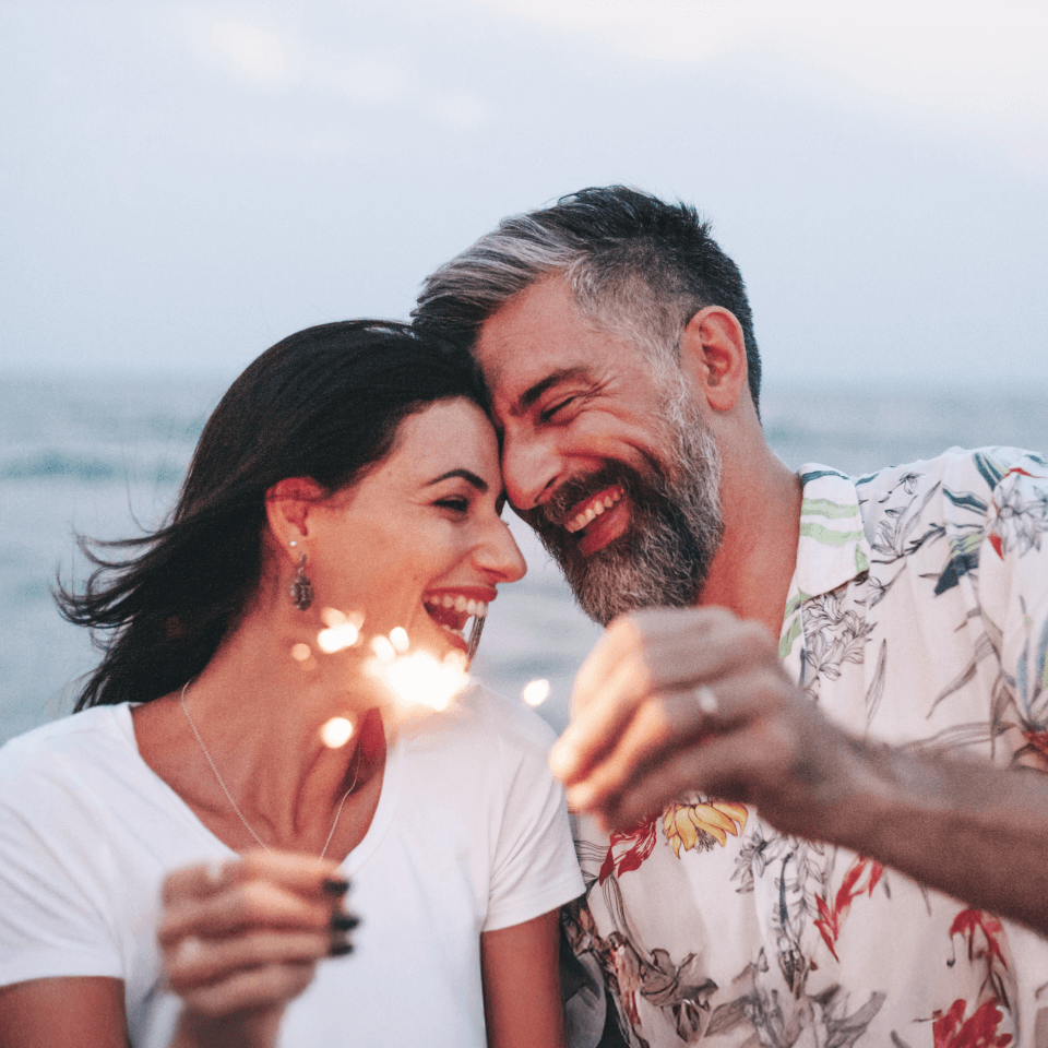 A couple laughing while playing with sparklers near the shore.