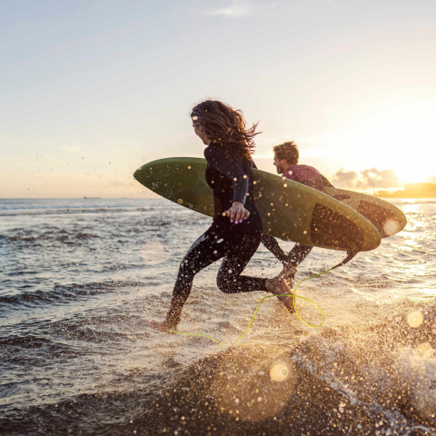 Two people in wetsuits running into the waves with their surfboards as the sun sets behind them.