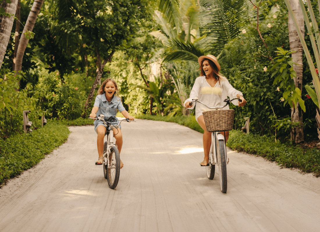 A woman and girl joyfully bike together through a sandy path surrounded by green foliage.