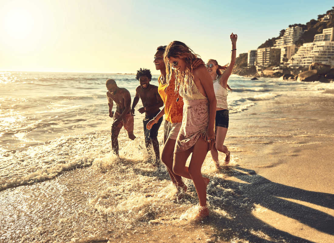 A group of five smiling friends walking along the beach on a bright morning and splashing around.