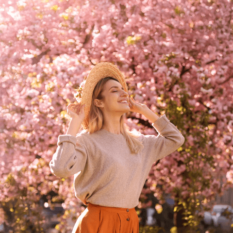 A smiling lady with a straw hat standing in front of beautiful, blooming cherry blossoms.