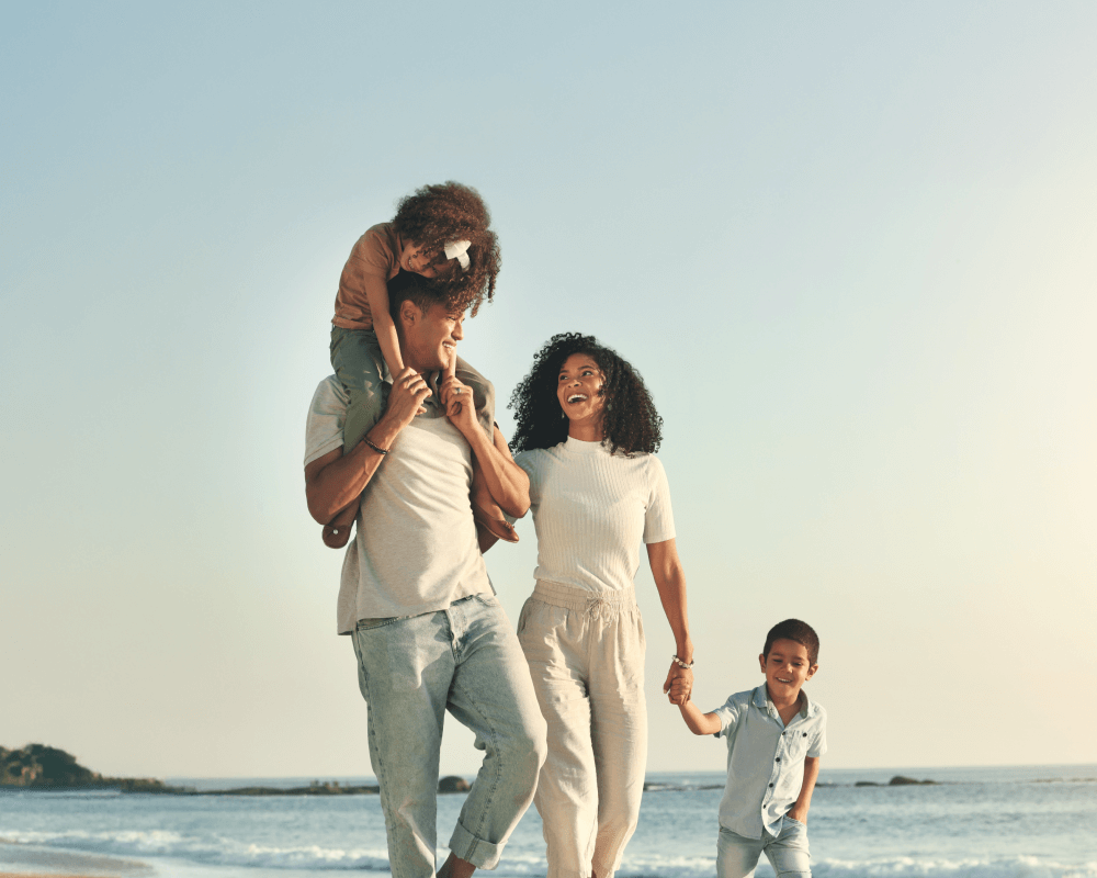 A smiling dad carrying his daughter on his shoulders while walking with his smiling wife and happy son along a beach in the early evening.