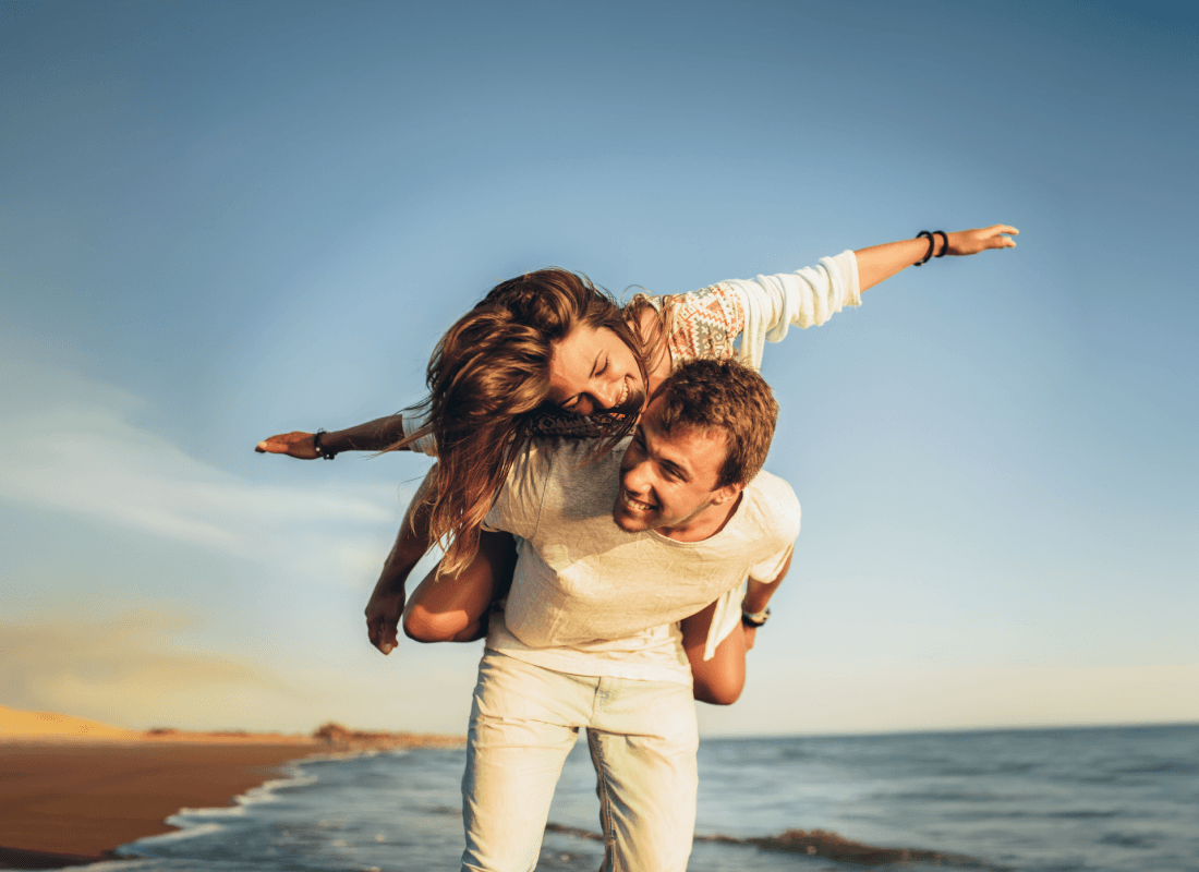A man piggybacking a lady with her arms outstretched while they walk along an empty beach at dusk.