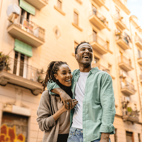A happy couple walking arm-in-arm along a European city street with historic apartment buildings in the background.
