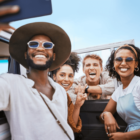 A group of four smiling friends taking a selfie together while hanging out around an open-window vehicle.