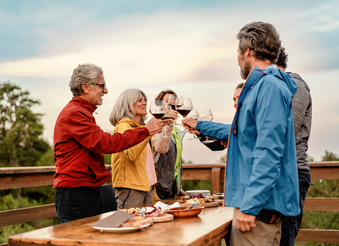 Six people standing outside around a wooden table with various plates, while they toast with red wine.
