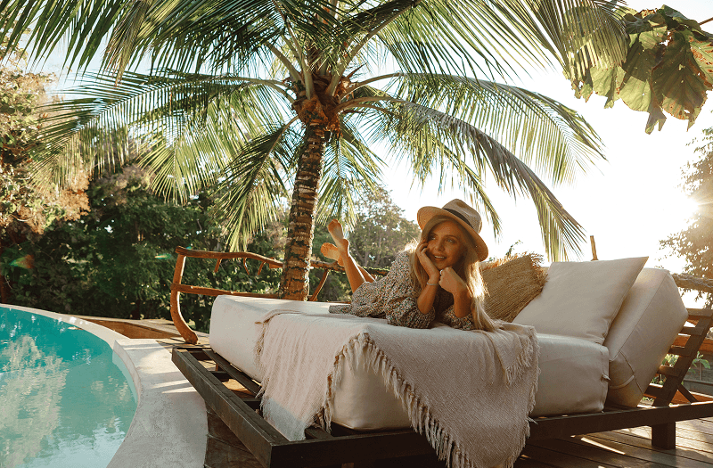 A woman with a hat stretched out on a white lounger underneath a tall palm tree beside a resort pool.