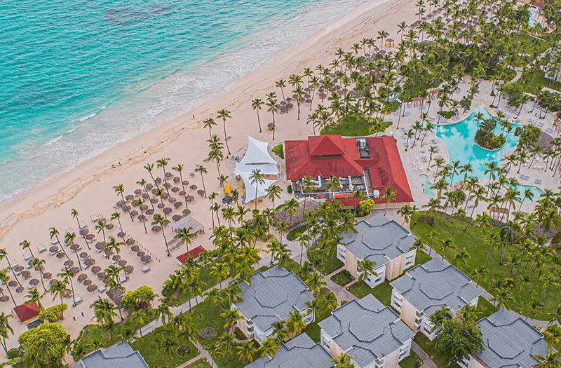 Aerial view of Grand Bavaro Princess All Suites Resort, Spa & Casino in Punta Cana with a bright red building, expansive pool, and many palm umbrellas.