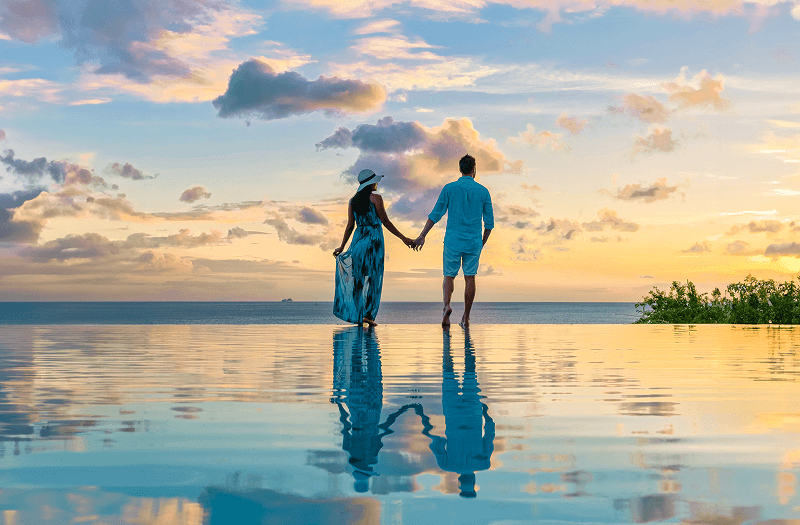 A woman in a long blue dress and a man in a blue shirt and shorts holding hands and standing on the beach watching a beautiful sunset.