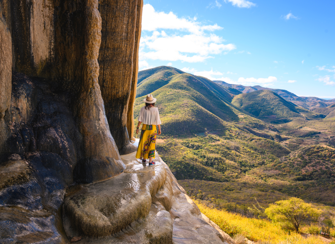 A woman in a hat and colorful skirt standing on the edge of a cliff face looking at mountains in the distance