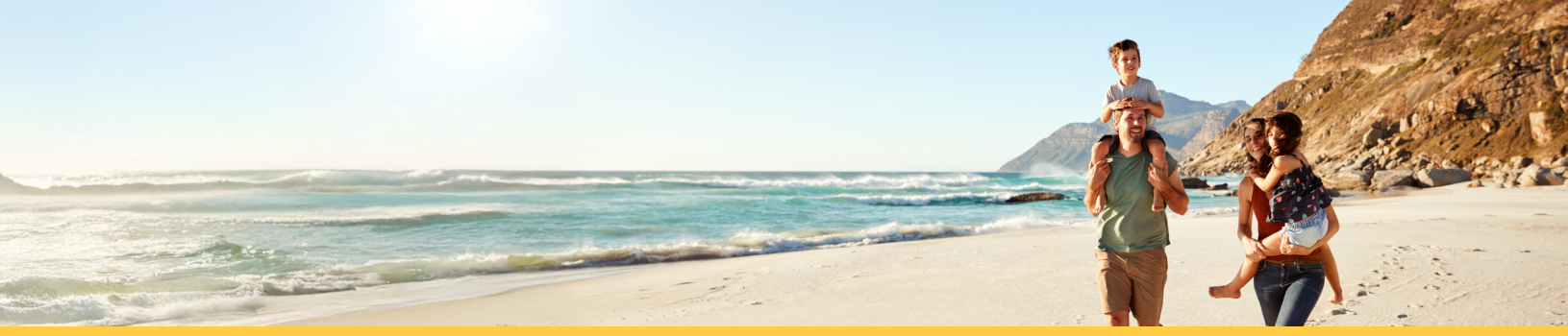 Two smiling parents holding their daughter and son while walking down a sandy beach away from a tall rock formation.