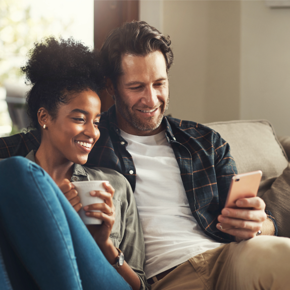 A smiling couple sitting on a couch and looking at a smartphone together.