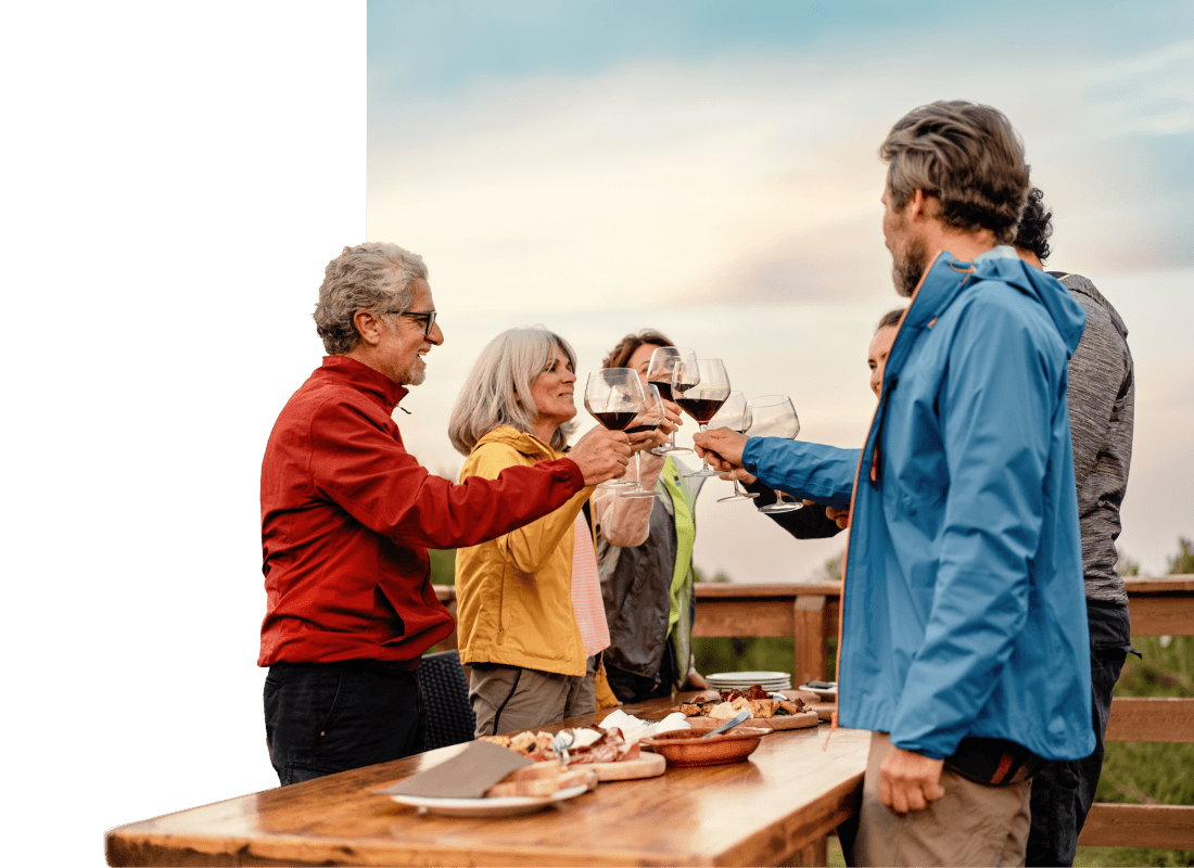 Six people standing outside around a wooden table with various plates, while they toast with red wine.