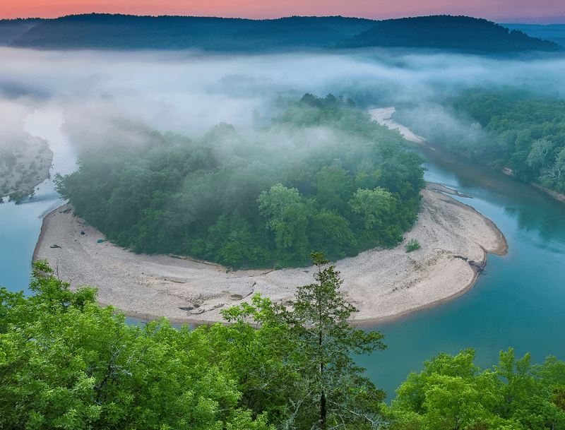 An aerial view of a horseshoe-shaped river bend in Arkansas surrounded by dense green forests, with a low layer of morning fog rolling over the trees and water.