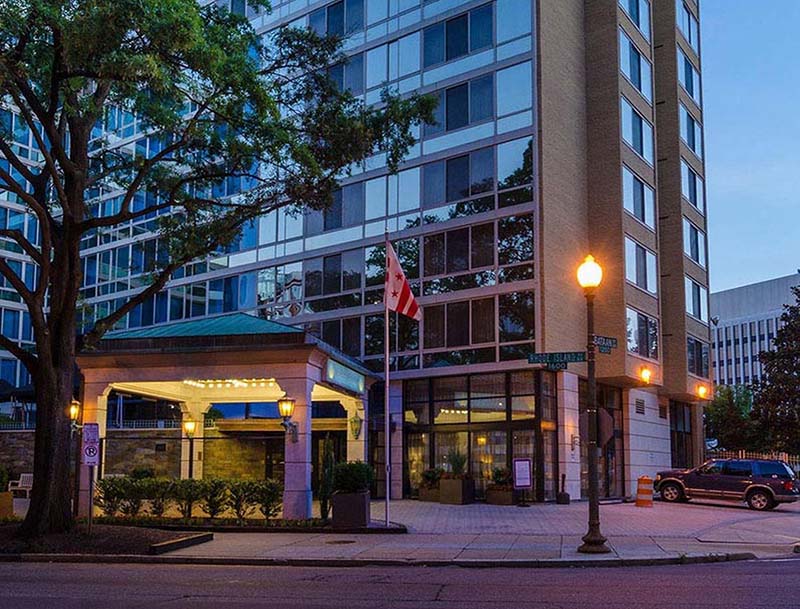 A dusk view of the hotel's illuminated entrance and facade with large windows, as seen from the street.