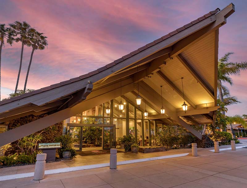 A dusk view of the hotel's entrance featuring a tropical-inspired, sloped roof, warm lighting, and lush landscaping.