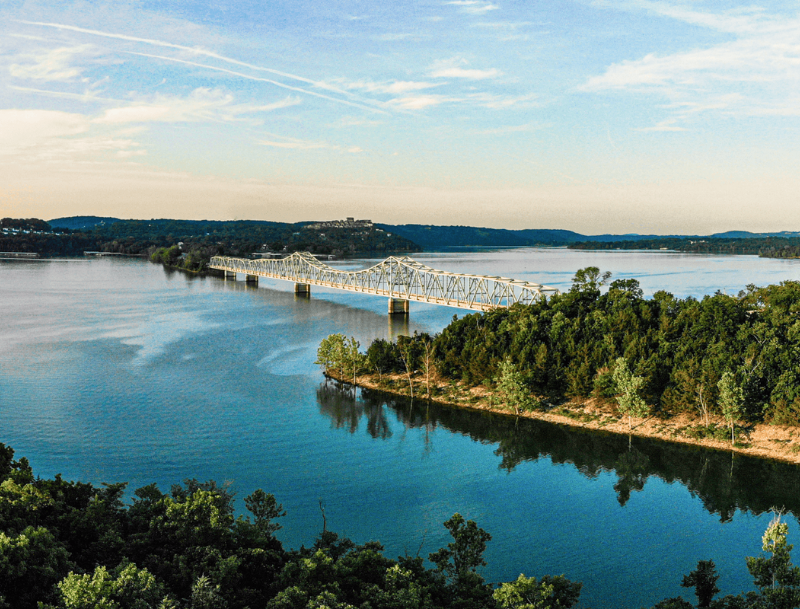 An aerial view of a long steel-truss bridge spanning across Lake Taneycomo in Branson, Missouri, surrounded by lush green forested shorelines under a hazy sky.