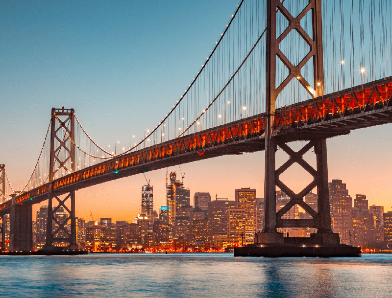 The illuminated Bay Bridge suspension towers and cables at dusk, with the glowing San Francisco skyline across the water.