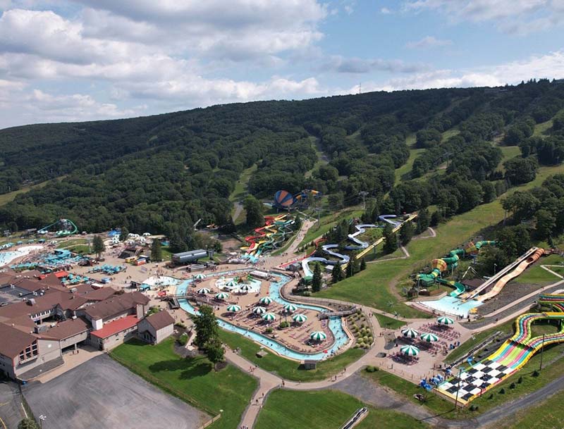 Aerial view of a vibrant hillside water park featuring a lazy river, pools, water slides and other attractions surrounded by lush natural greenery.