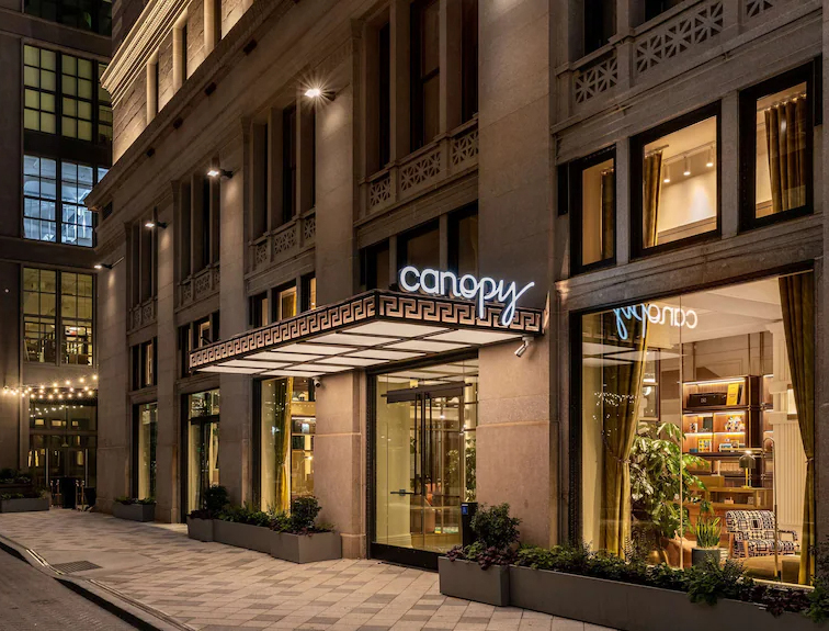 The hotel’s modern entrance at night, featuring a decorative geometric awning with illuminated signage and large glass windows revealing a warm lobby interior.