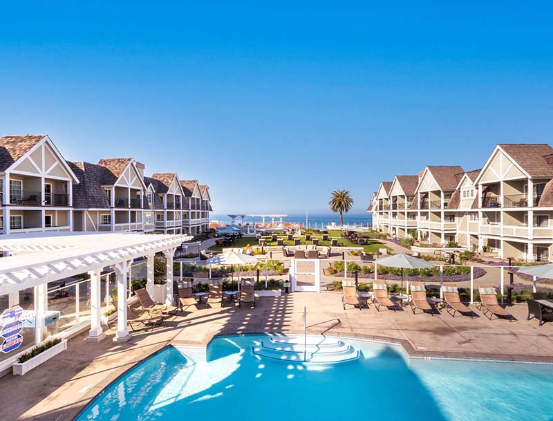 A view of the pool and deck area nestled between two rows of resort buildings, with the ocean visible in the background.