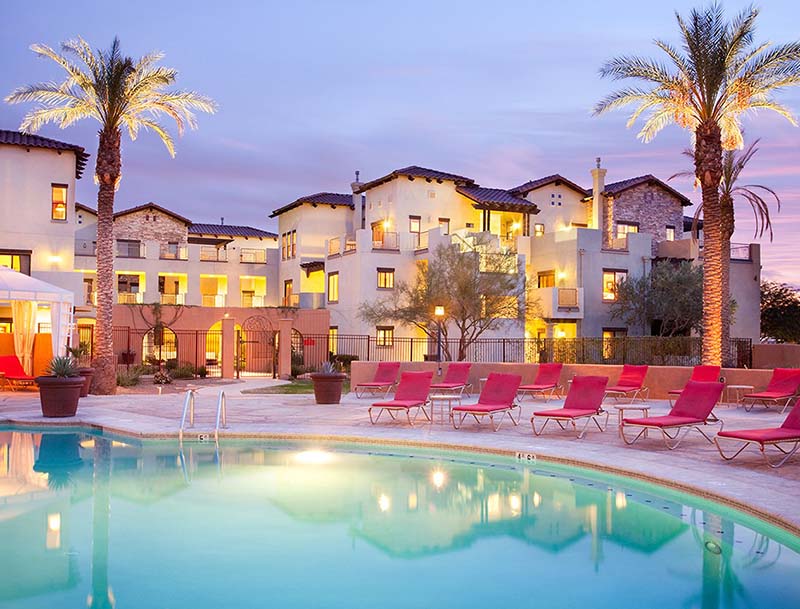 The resort pool at sunset, surrounded by lounge chairs and palm trees, with the brightly lit resort buildings visible in the background. 