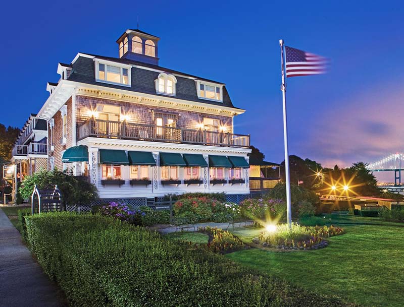 A dusk view of Bay Voyage Inn's illuminated facade and lush landscaping, with the Claiborne Pell Newport Bridge visible in the distance.