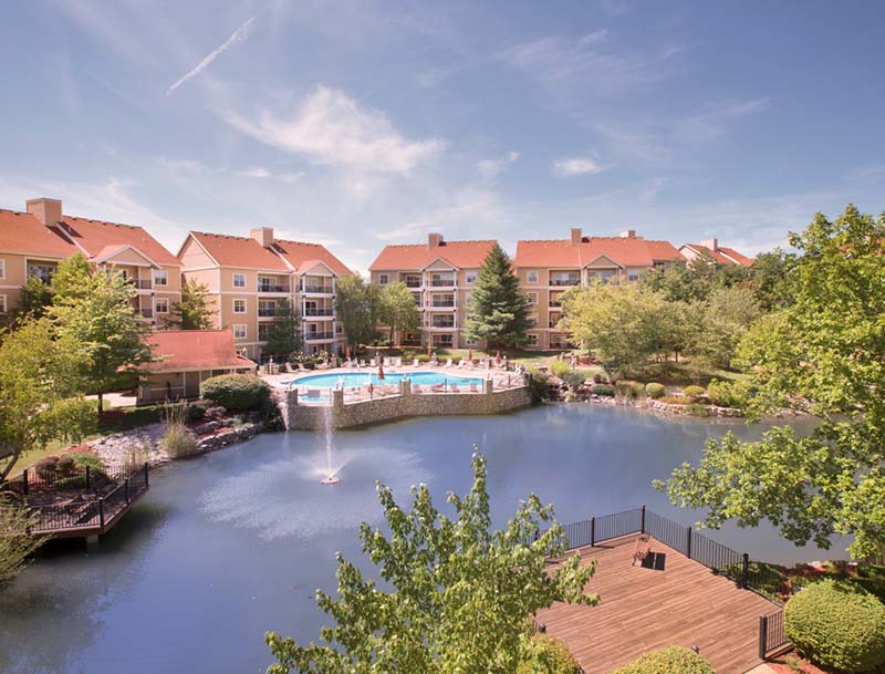 A bird's-eye view of a lake featuring sundecks and lush landscaping, with the resort buildings and pool visible in the background. 