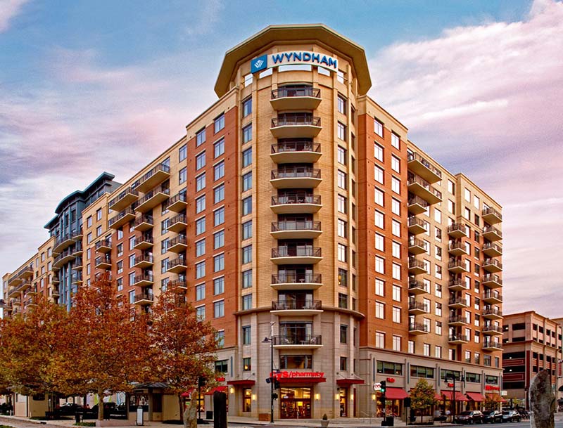 The hotel's welcoming entrance and brightly-colored facade with large windows and balconies, as seen from the tree-lined street.