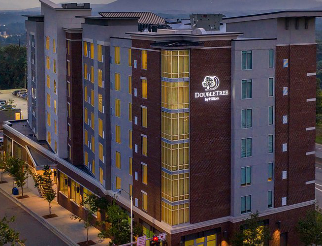 The hotel’s multi-story modern facade at dusk, featuring illuminated windows and prominent glowing signage, with a view of the Blue Ridge Mountains in the distance.