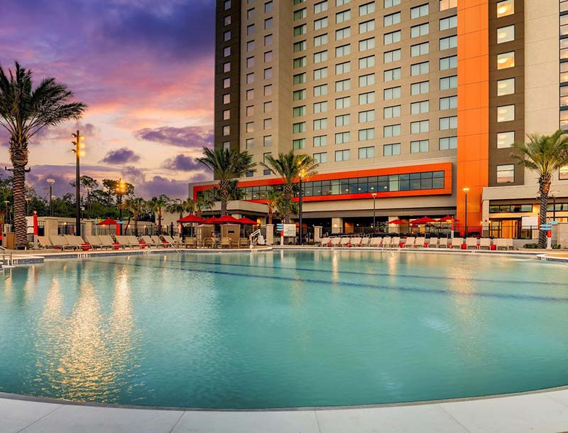 A sunset view of a large serene pool surrounded by lounge chairs and umbrellas, with the hotel's colorful facade in the background.